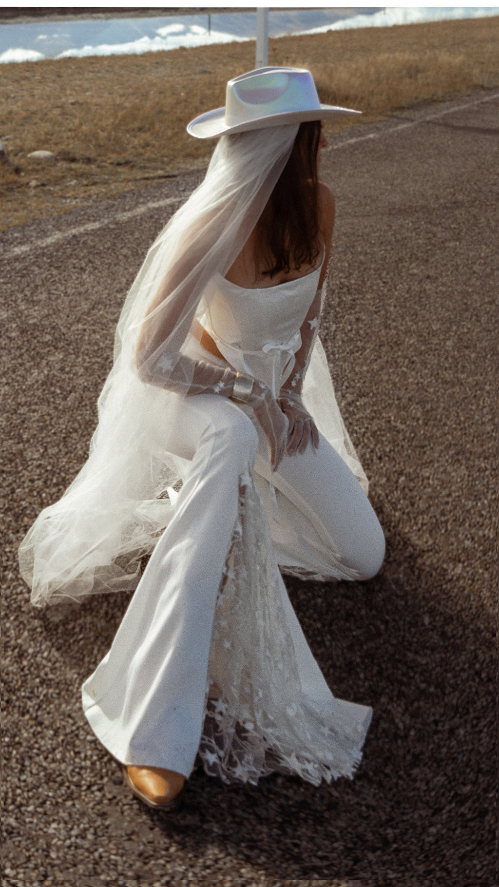 Kneeling pose in flared crepe bridal trousers with star lace insets, cowboy hat and long veil, road and lake behind, Sun Flares