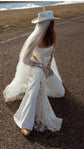 Kneeling pose in flared crepe bridal trousers with star lace insets, cowboy hat and long veil, road and lake behind, Sun Flares