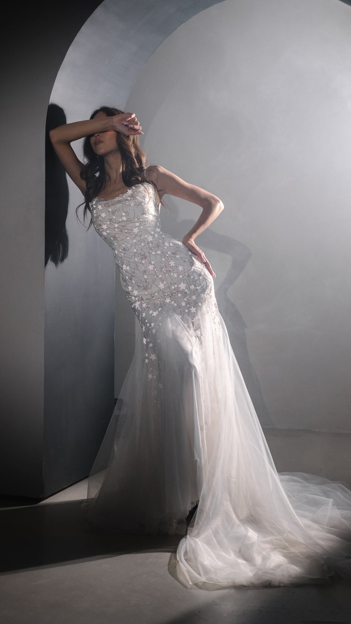 Full-length star beaded wedding dress with a flared tulle train, model leaning in an arched doorway, Supernova
