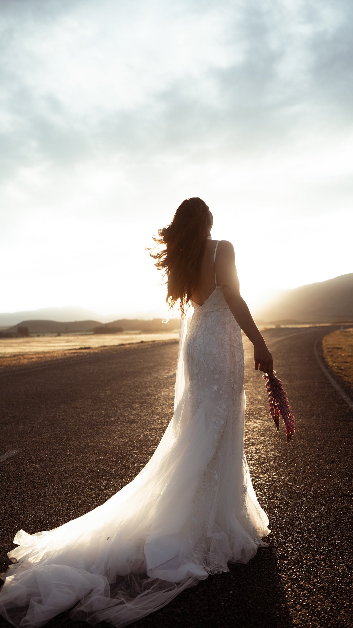 Bride walking down an open road at sunset in a low-back star wedding dress, tulle train and lupins in hand, elopement, Supernova
