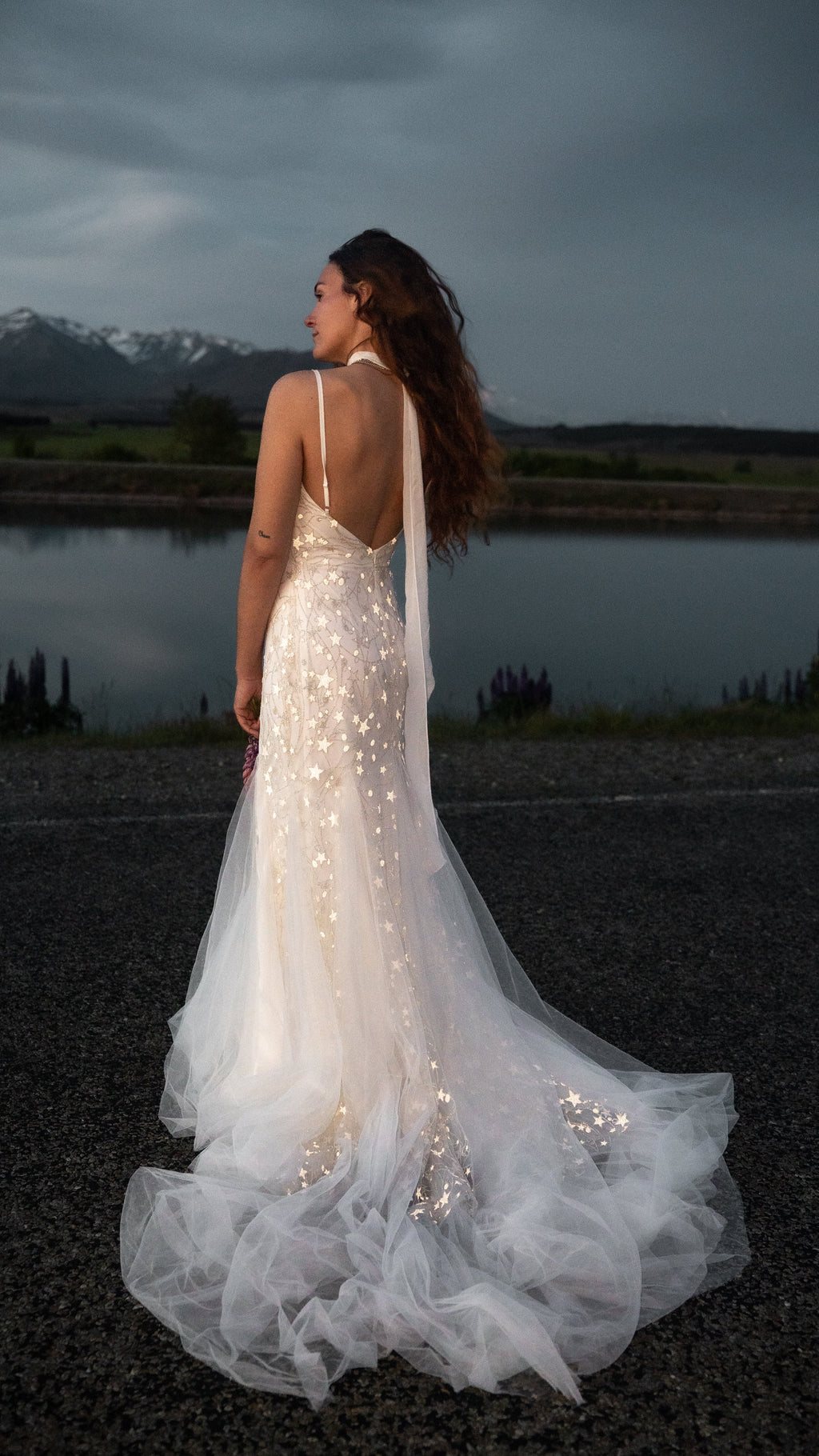 Bride by a lake at dusk in a starry fitted wedding dress with an open back, tulle train spread on the gravel, Supernova