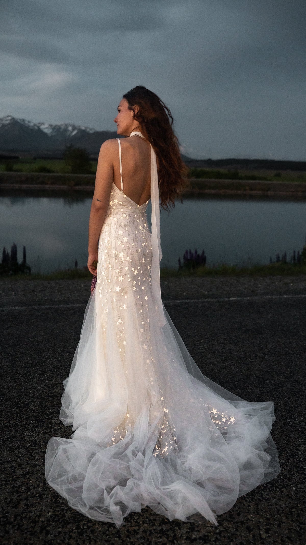 Bride by a lake at dusk in a starry fitted wedding dress with an open back, tulle train spread on the gravel, Supernova