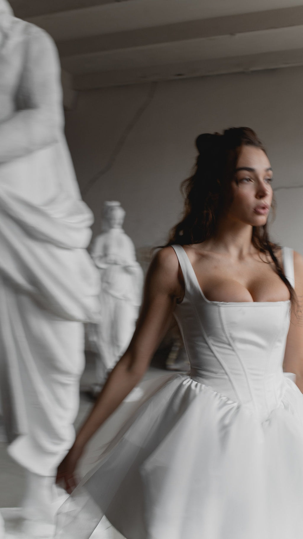 Satin corset mini wedding dress with a square neck, model glancing sideways among white statues, editorial, Swan