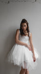 Model leaning on a pale wall in a short satin corset wedding dress with an uneven layered tulle hem, Swan