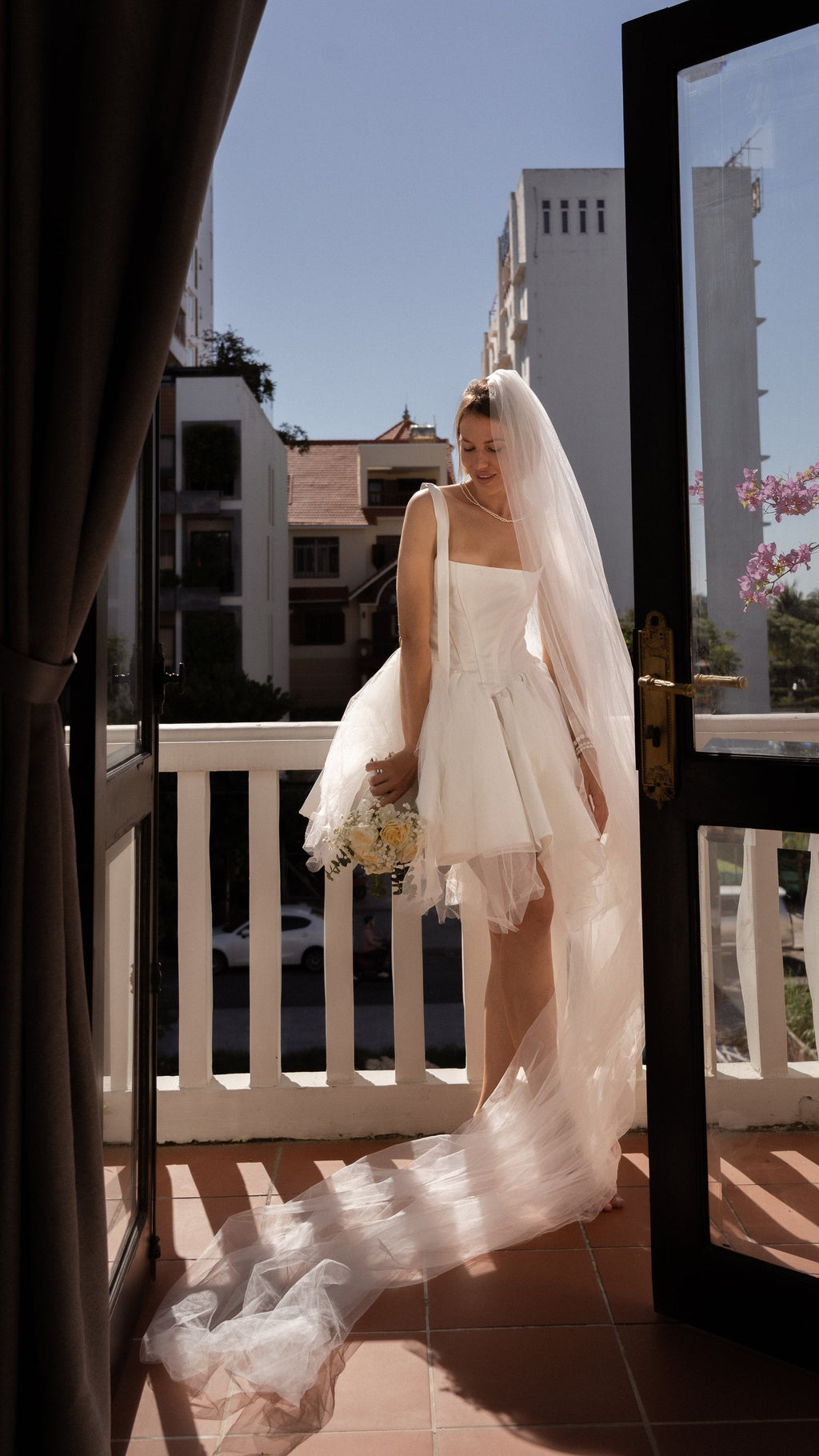 Bride on a sunny balcony in a corset mini wedding dress with a long tulle veil trailing over the tiles, bouquet, Swan