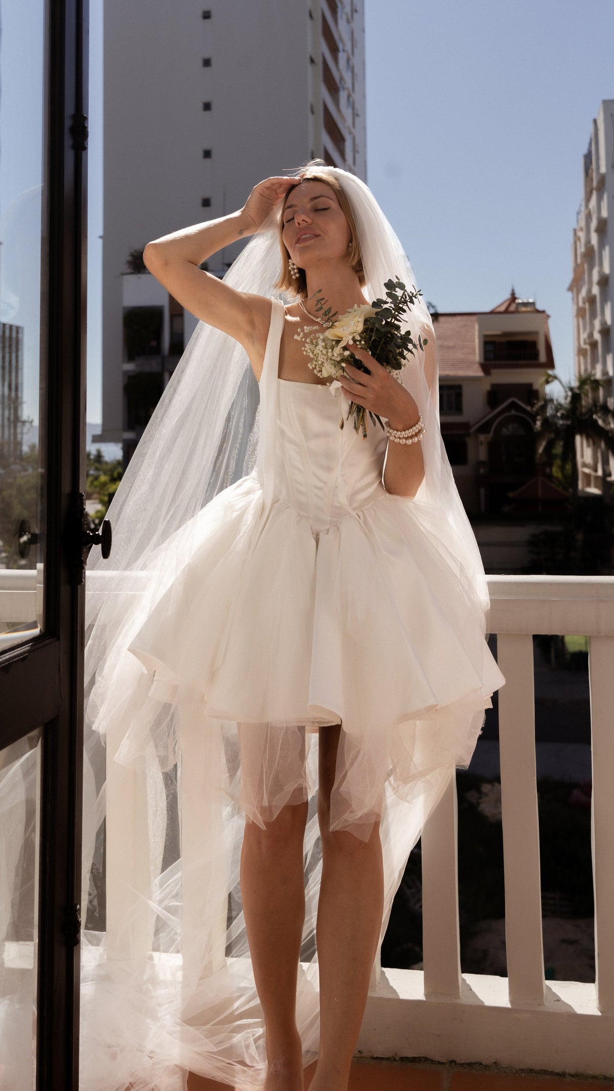 Bride with eyes closed in the sun in a square-neck corset mini wedding dress, long veil and white bouquet, Swan