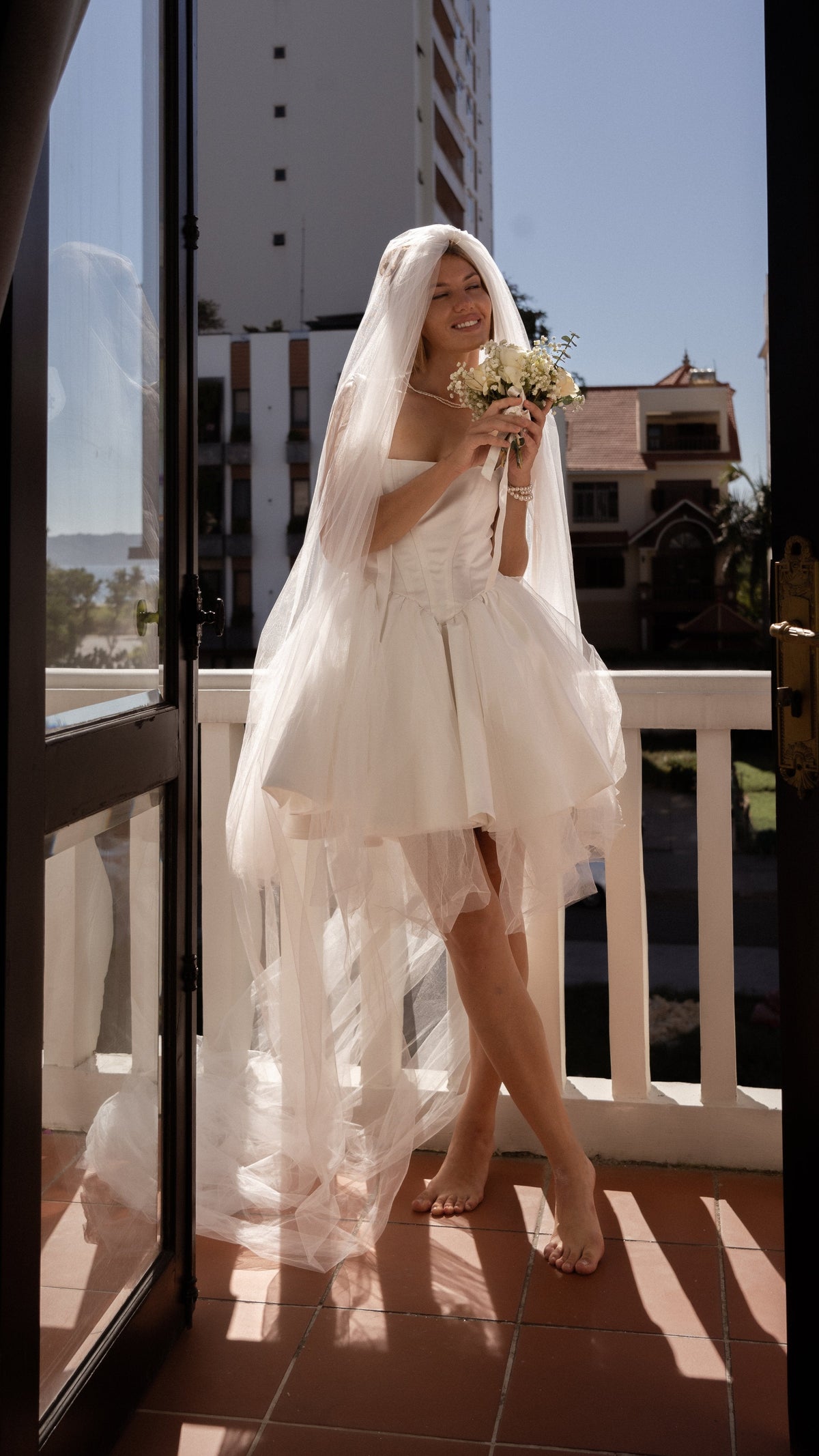 Barefoot bride smiling under a long veil on a balcony, short satin corset wedding dress and a small bouquet, Swan