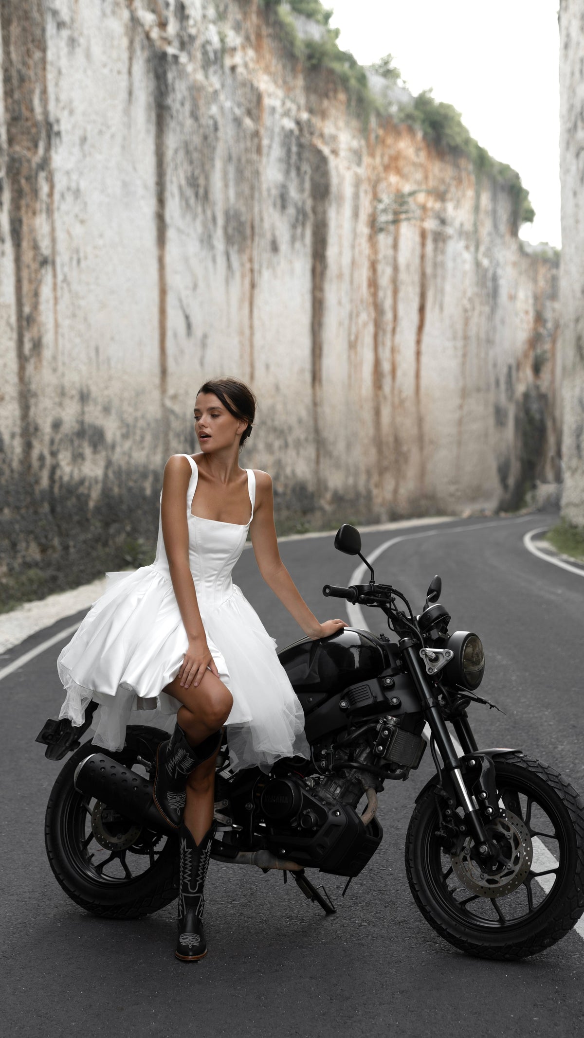 Bride perched on a motorbike in a square-neck corset wedding dress with a tulle mini skirt, rock walls behind, Swan