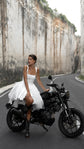 Bride perched on a motorbike in a square-neck corset wedding dress with a tulle mini skirt, rock walls behind, Swan