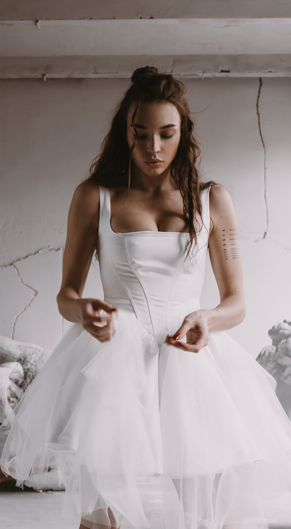 Model in a square-neck satin corset wedding dress with a dropped basque waist and tulle mini skirt, plaster wall, Swan