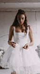 Model in a square-neck satin corset wedding dress with a dropped basque waist and tulle mini skirt, plaster wall, Swan