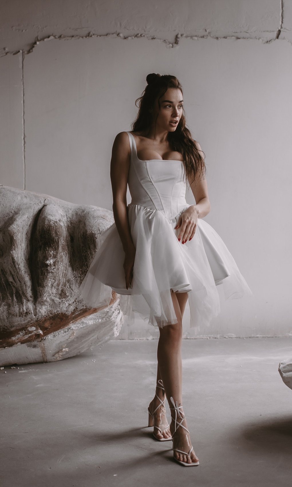 Model stepping forward in a short corset wedding dress with a basque waist and tulle skirt, plaster sculpture behind, Swan