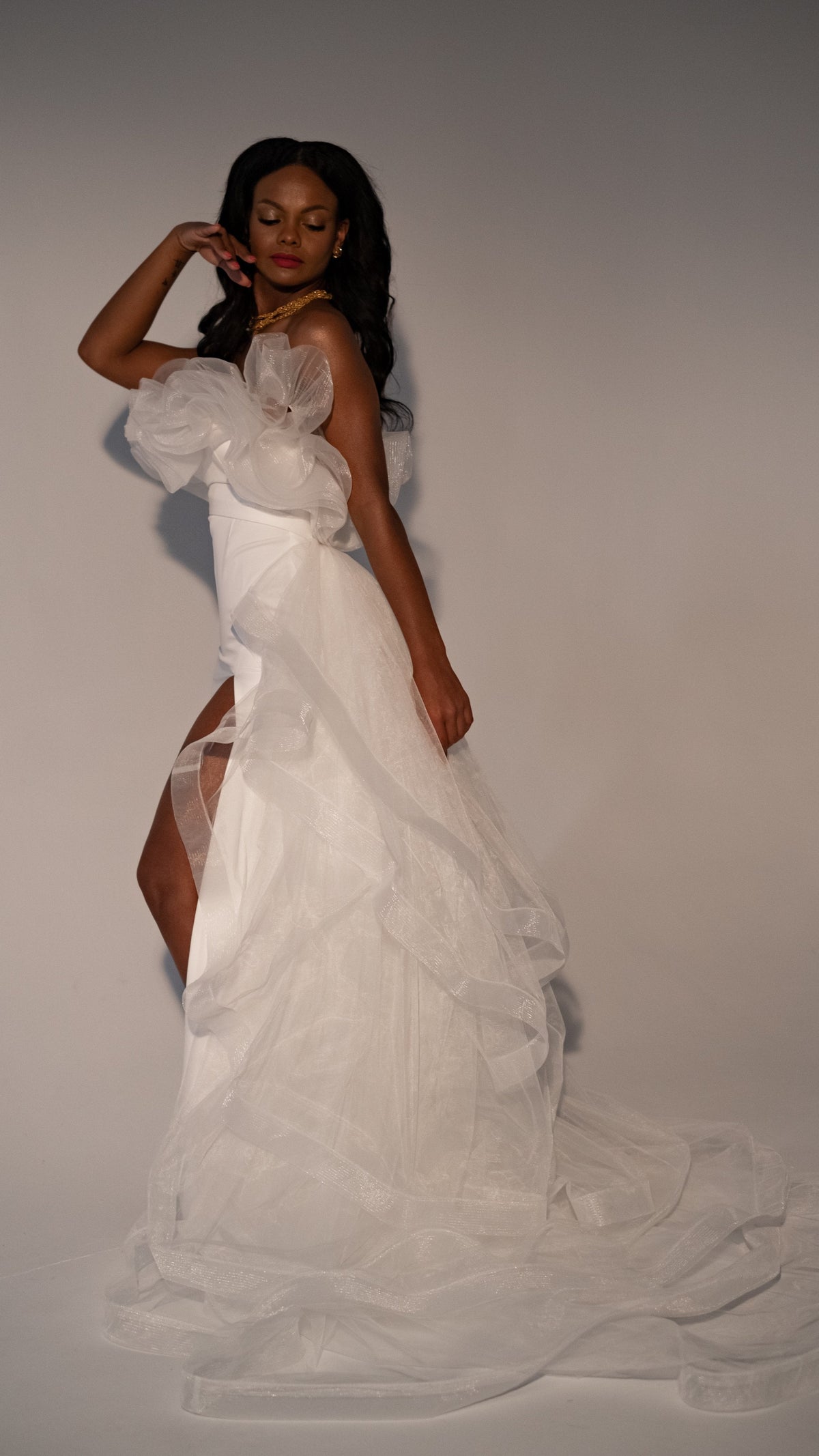 Model touching her hair in a fitted crepe sample wedding dress with a tiered tulle overskirt to the floor, Terra Sample