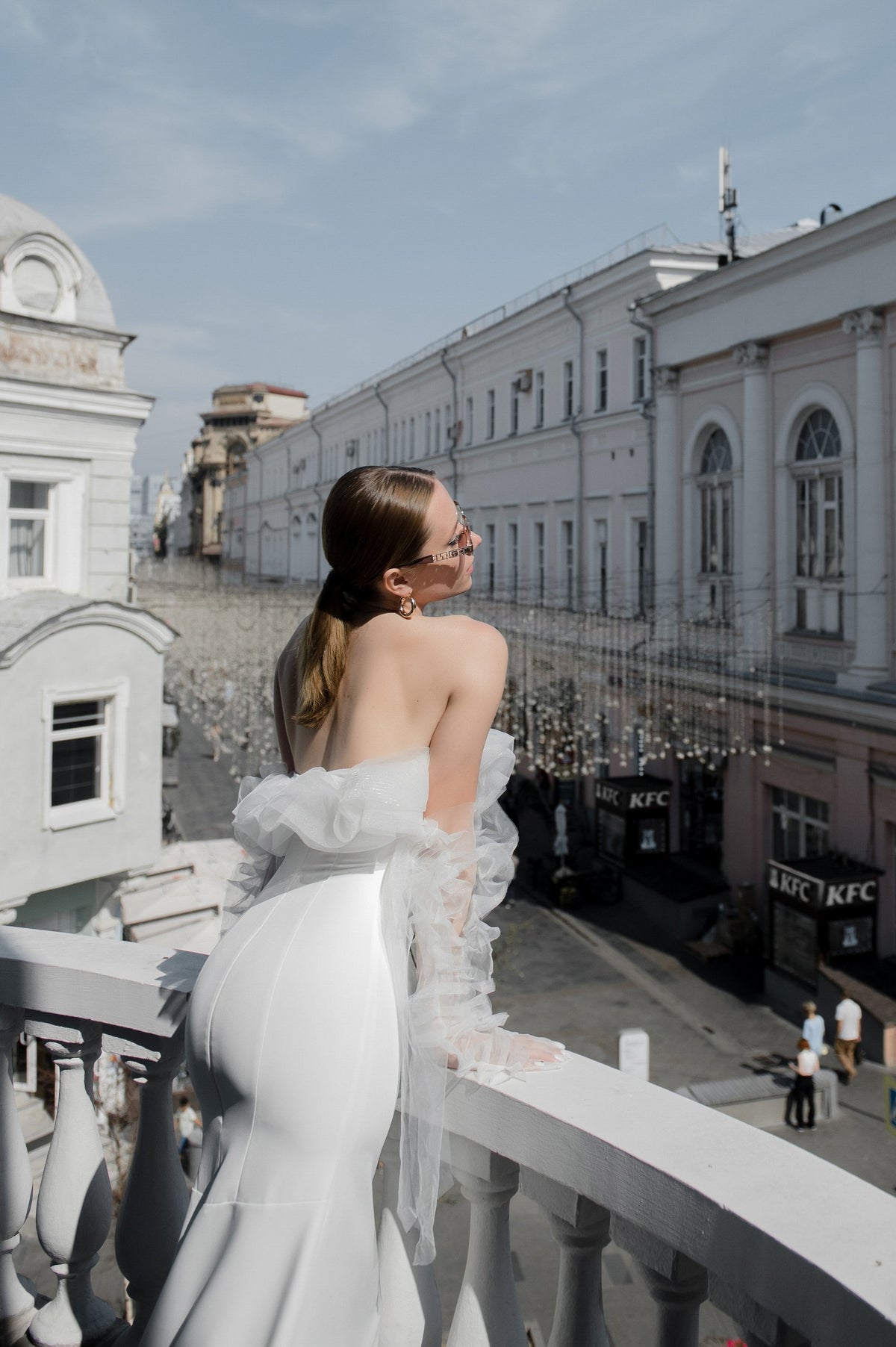 Back view of a crepe sample wedding dress on a city balcony, ruffled tulle at the shoulders and street below, Terra Sample
