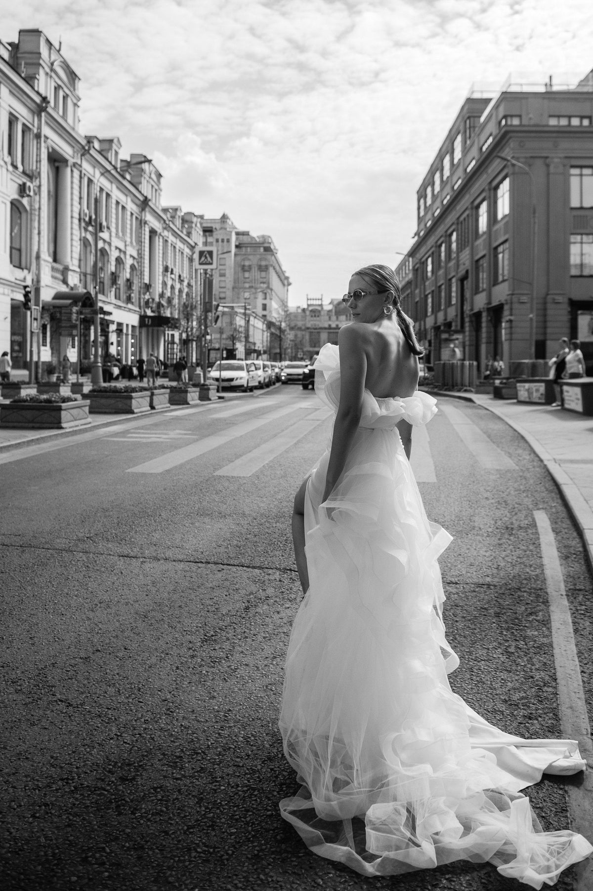 Black and white back view of a bride on a city street in a sample wedding dress with a ruffled tulle train, Terra Sample