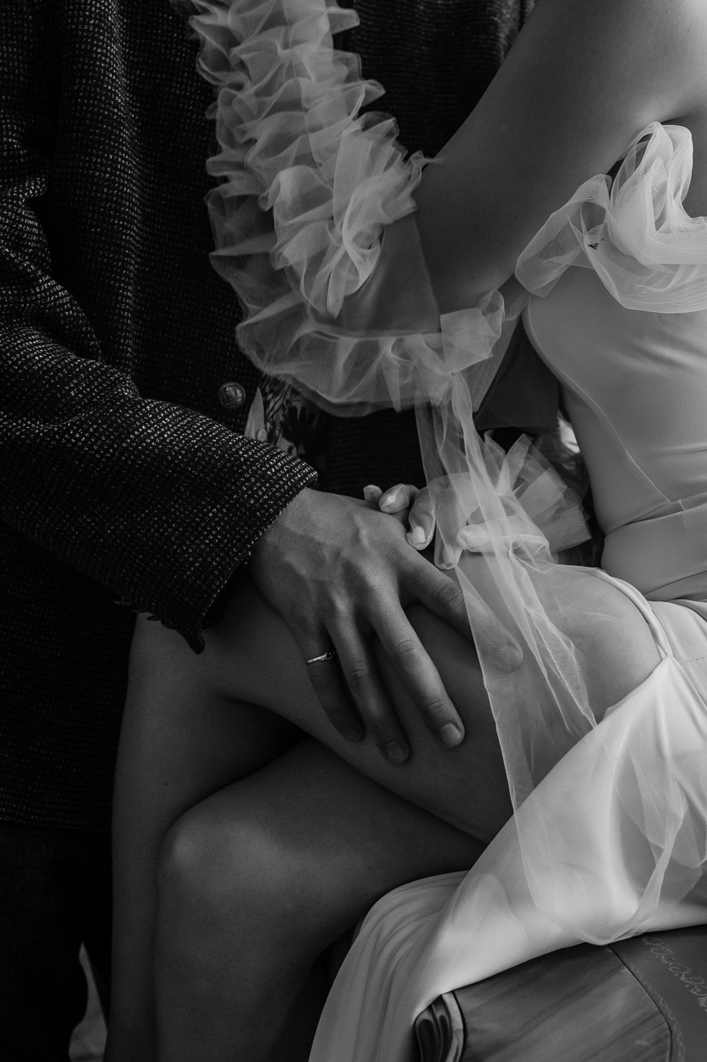 Black and white close-up of the ruffled tulle shoulder of a crepe sample wedding dress, groom's hand on her knee, Terra Sample