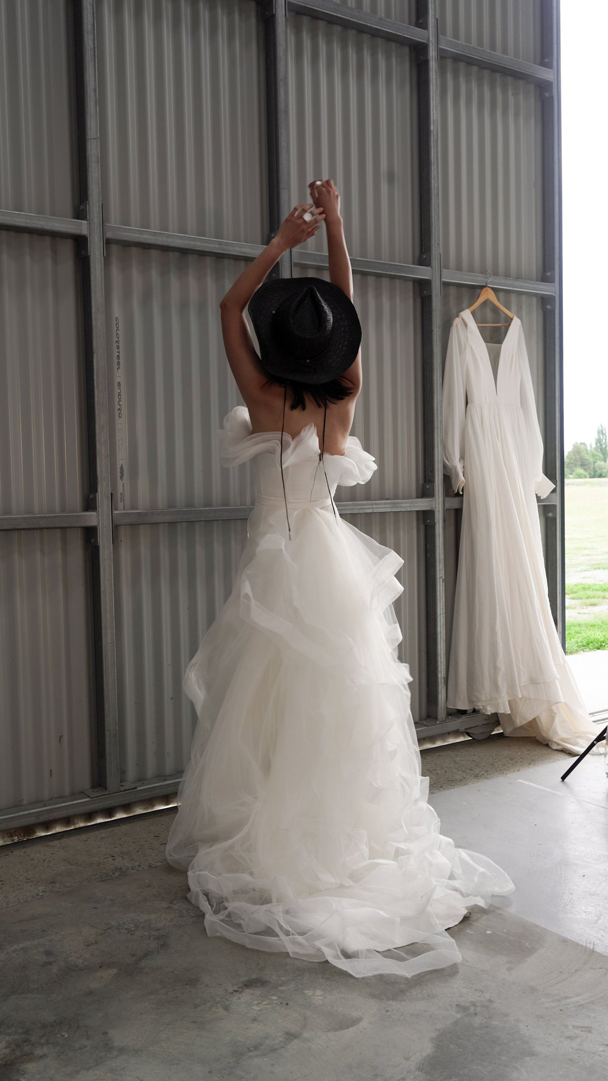 Bride with arms raised holding a hat, ruffled off-shoulder sample wedding dress with a tiered tulle train, Terra Sample