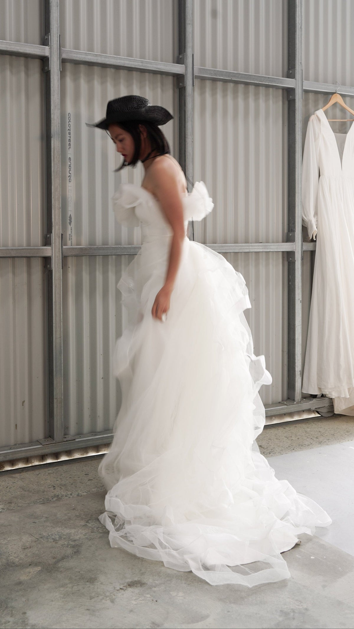 Bride in a black hat gathering the tulle overskirt of a crepe sample wedding dress, metal shed with a hanging gown, Terra Sample