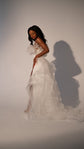 Model in a crepe sample wedding dress with the removable tulle overskirt cascading into a train, studio wall, Terra Sample