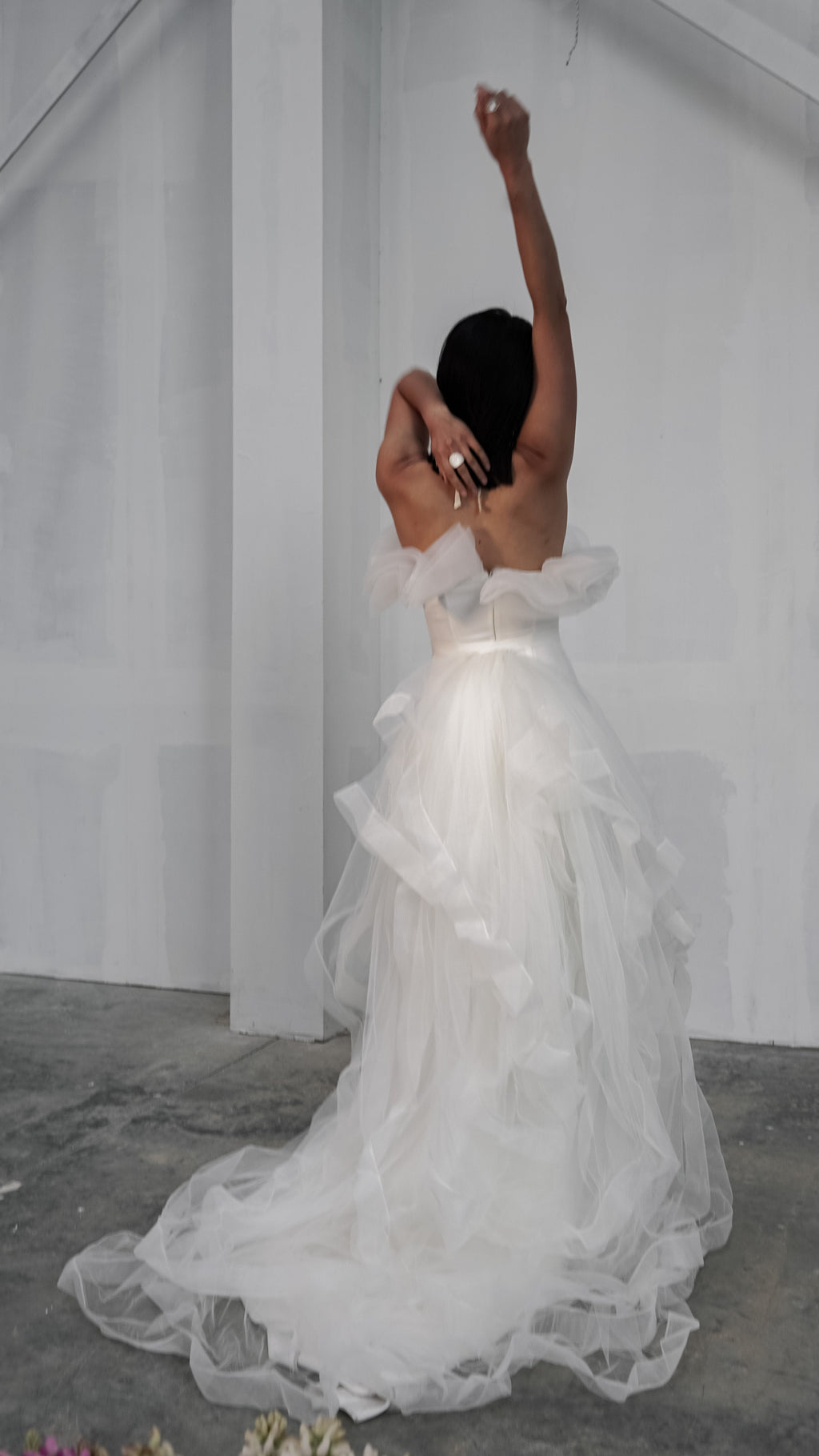 Back view of a bride with one arm raised, off-shoulder wedding dress and ruffled tulle overskirt trailing, Terra