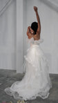 Back view of a bride with one arm raised, off-shoulder wedding dress and ruffled tulle overskirt trailing, Terra