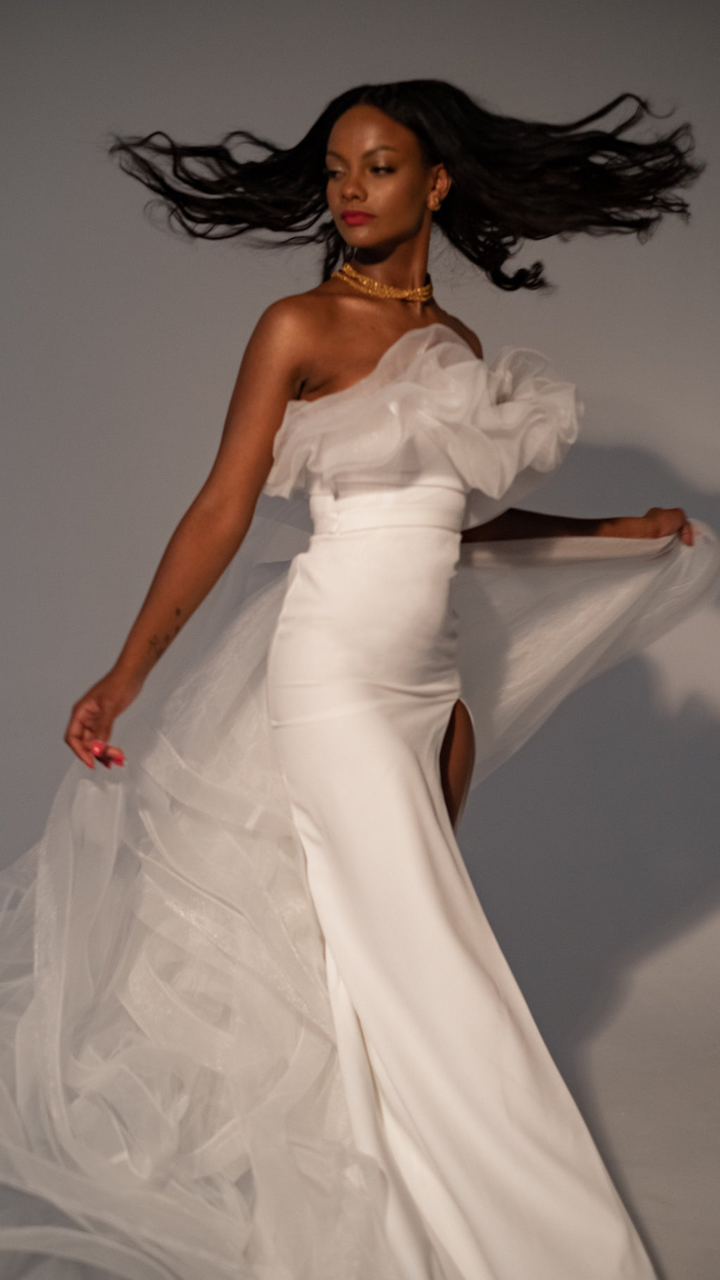 Model spinning in a crepe wedding dress with a tulle overskirt flying out, hair in motion against a grey wall, Terra