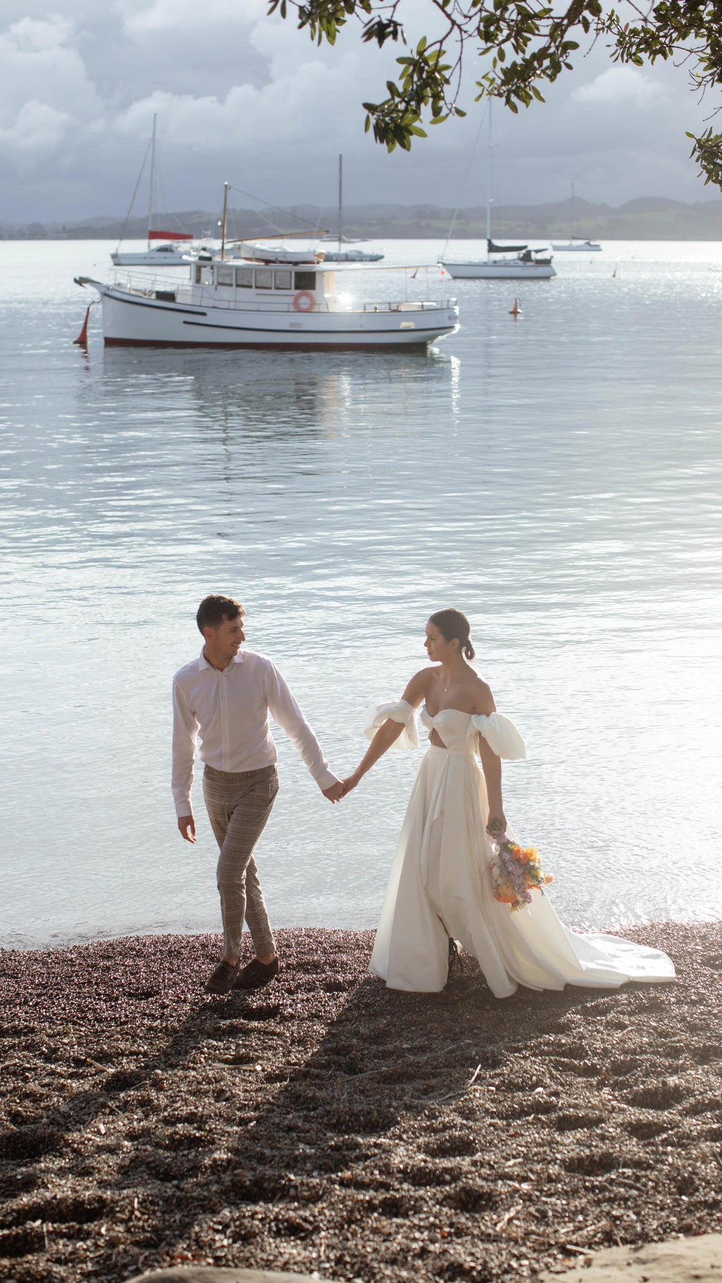 Bride and groom walking hand in hand on a pebble beach, satin wedding dress with a slit and puff sleeves, boats behind, The Dark Side of The Moon