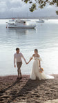 Bride and groom walking hand in hand on a pebble beach, satin wedding dress with a slit and puff sleeves, boats behind, The Dark Side of The Moon