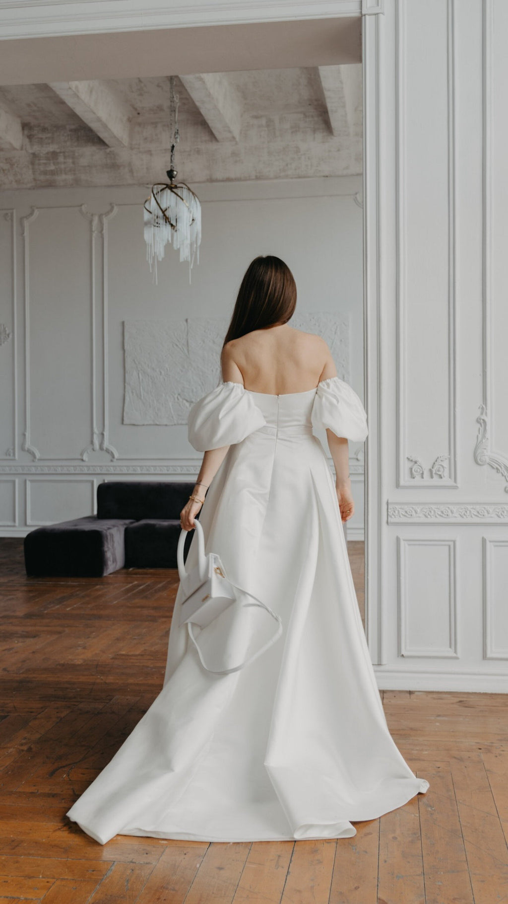 Back view of a bandeau satin wedding dress with detached puff sleeves and a long train, bride holding a white bag, The Dark Side of The Moon