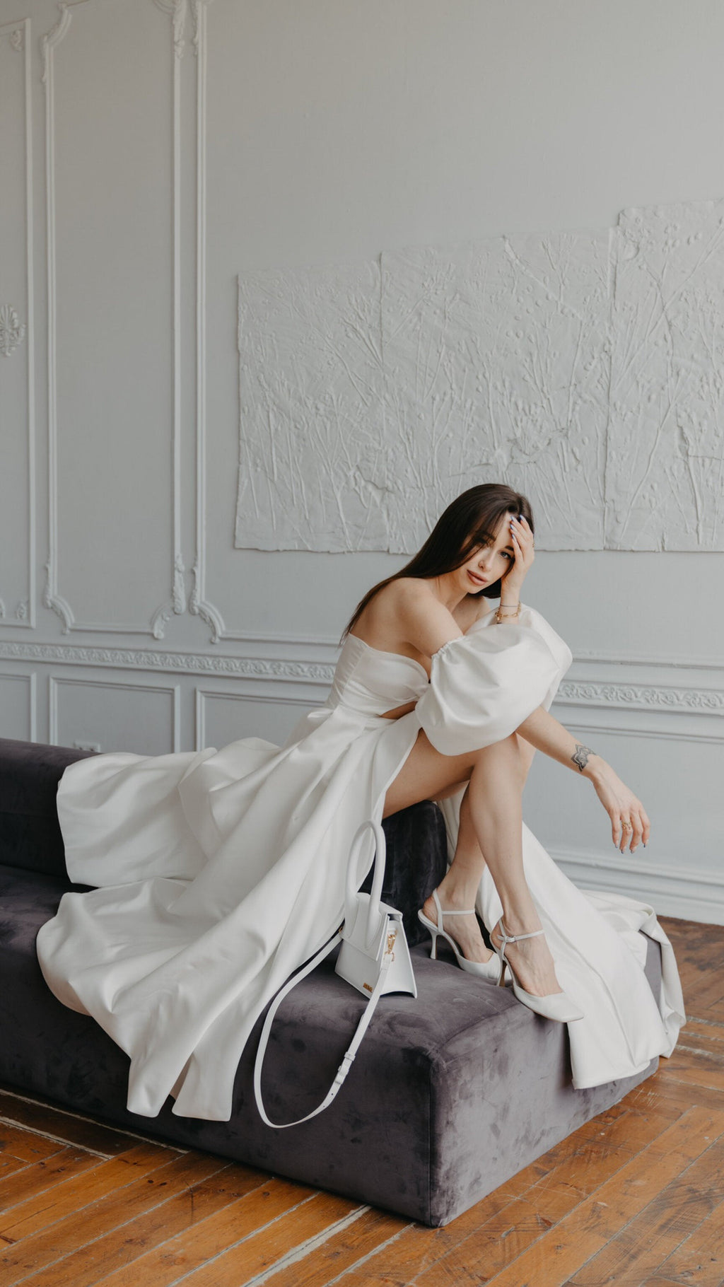 Bride seated on a velvet bench in a satin wedding dress with a wrap-front slit and puff sleeves, hand to her head, The Dark Side of The Moon