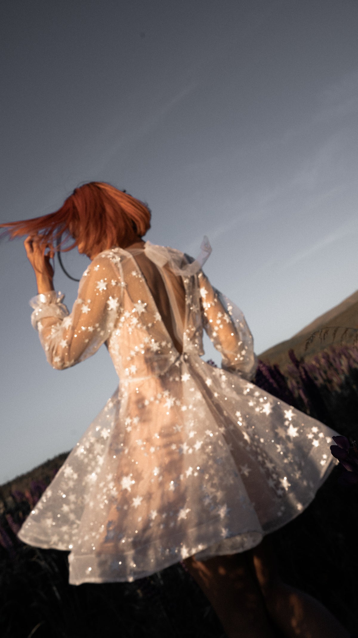 Celestial reception dress in beige star lace with long sheer sleeves and an open back, bride twirling in a lupin field, Twilight Beige by Ephemeris