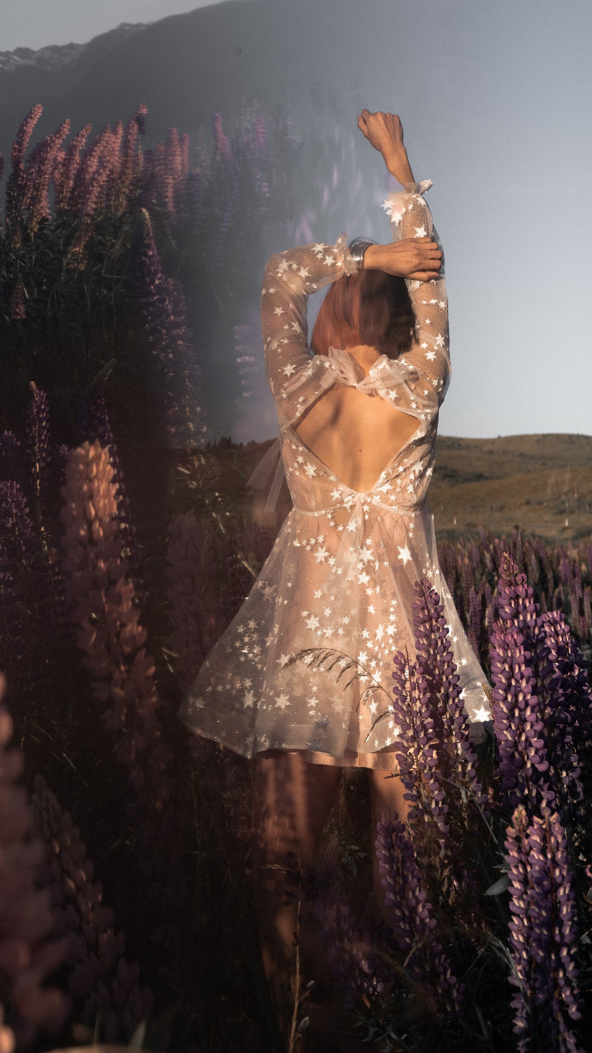 Back view of a short beige star reception dress with an open back and tie, arms raised among tall lupins, wildflower field, Twilight Beige