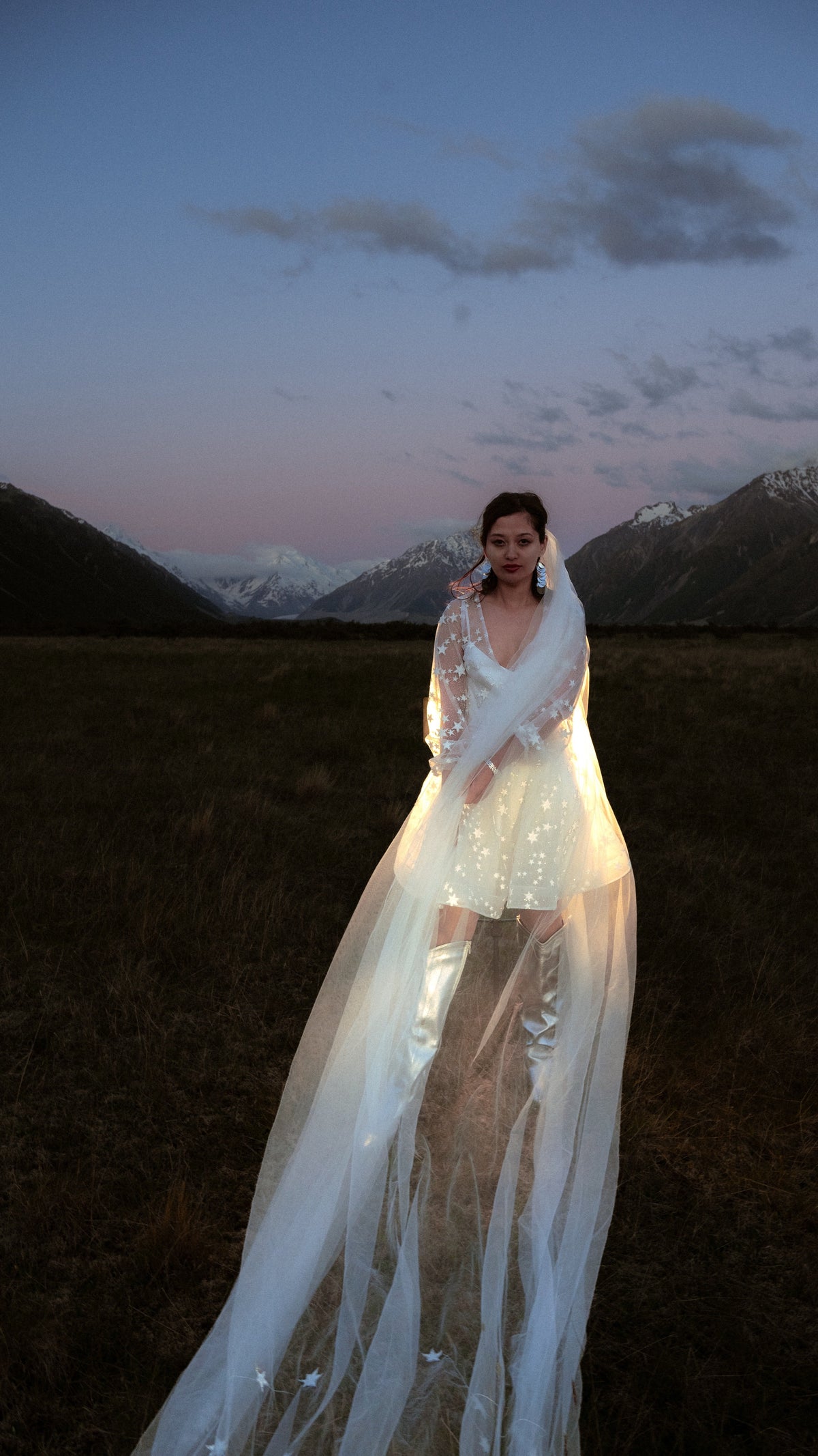 Bride in a celestial reception dress with sheer star sleeves under a long tulle veil, mountains at dusk behind, Twilight Ivory by Ephemeris