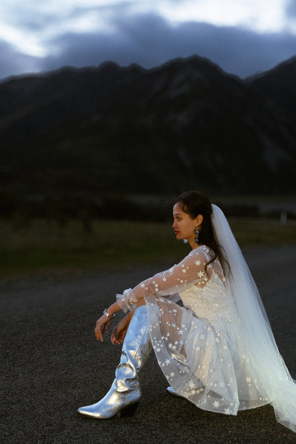 Bride sitting on the road in an ivory star reception dress with silver boots and a long veil, mountains behind, elopement, Twilight Ivory