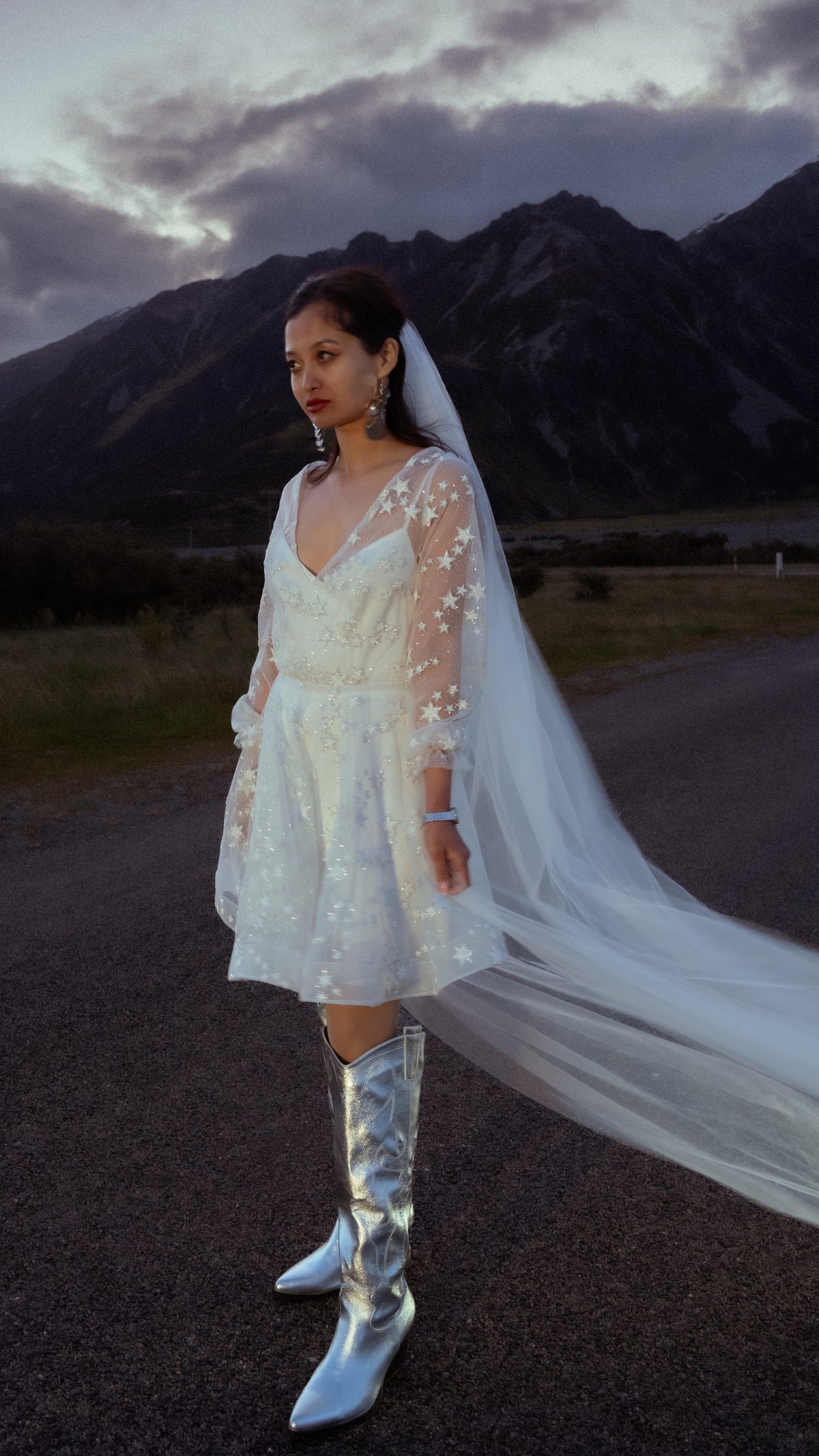Short ivory reception dress with star lace and sheer sleeves, bride glancing aside on a valley road, veil trailing, Twilight Ivory