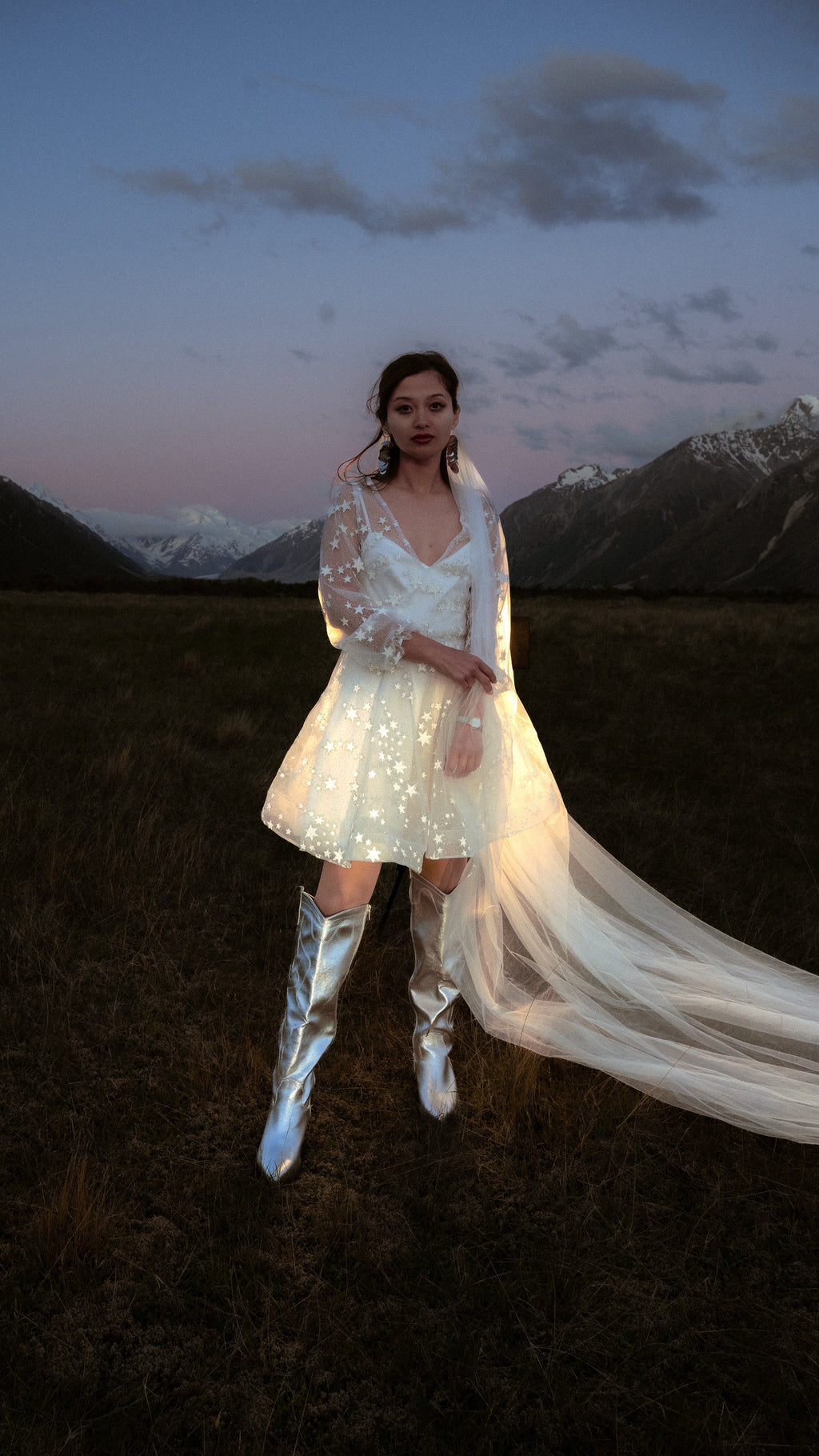 Ivory star reception dress lit by flash in a dusk field, bride with a long tulle veil and silver knee boots, Twilight Ivory