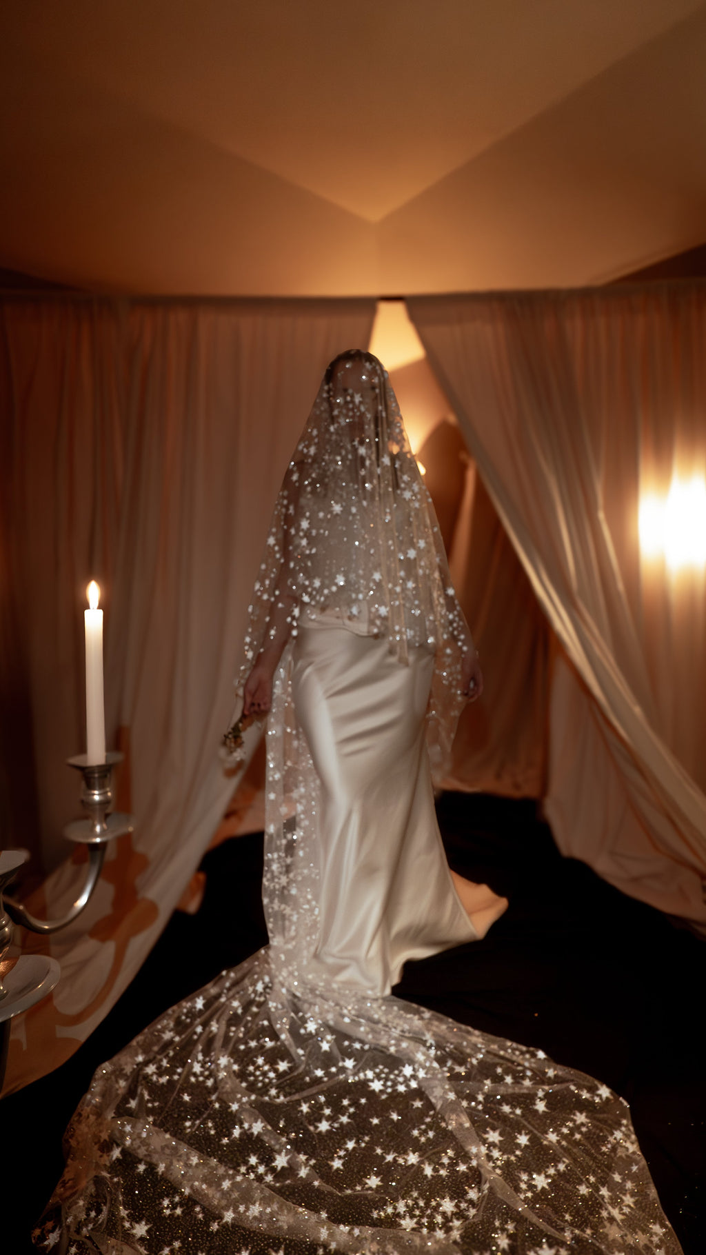 Bride turning beside a candelabra, star cathedral wedding veil worn over the face and pooling behind her, warm low light, Velum Veil