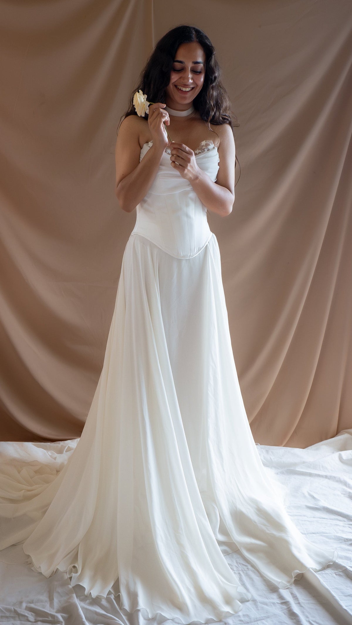Smiling bride holding a rose in a strapless bridal corset with a chiffon skirt spread on a white floor cloth, Venus Corset