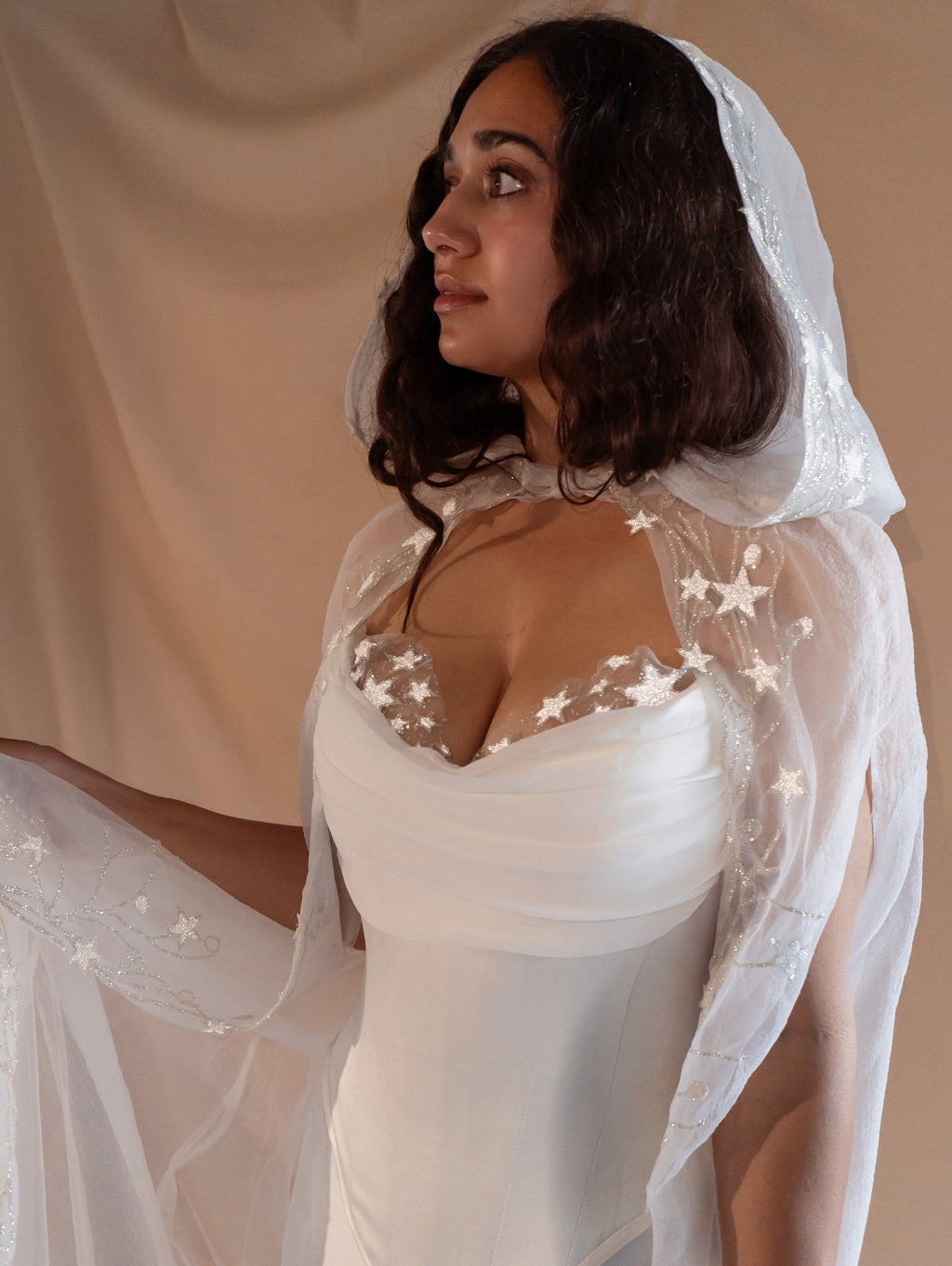 Profile of a bride in a hooded star cape over the satin corset of a bridal separates set, studio drape, Venus Ensemble