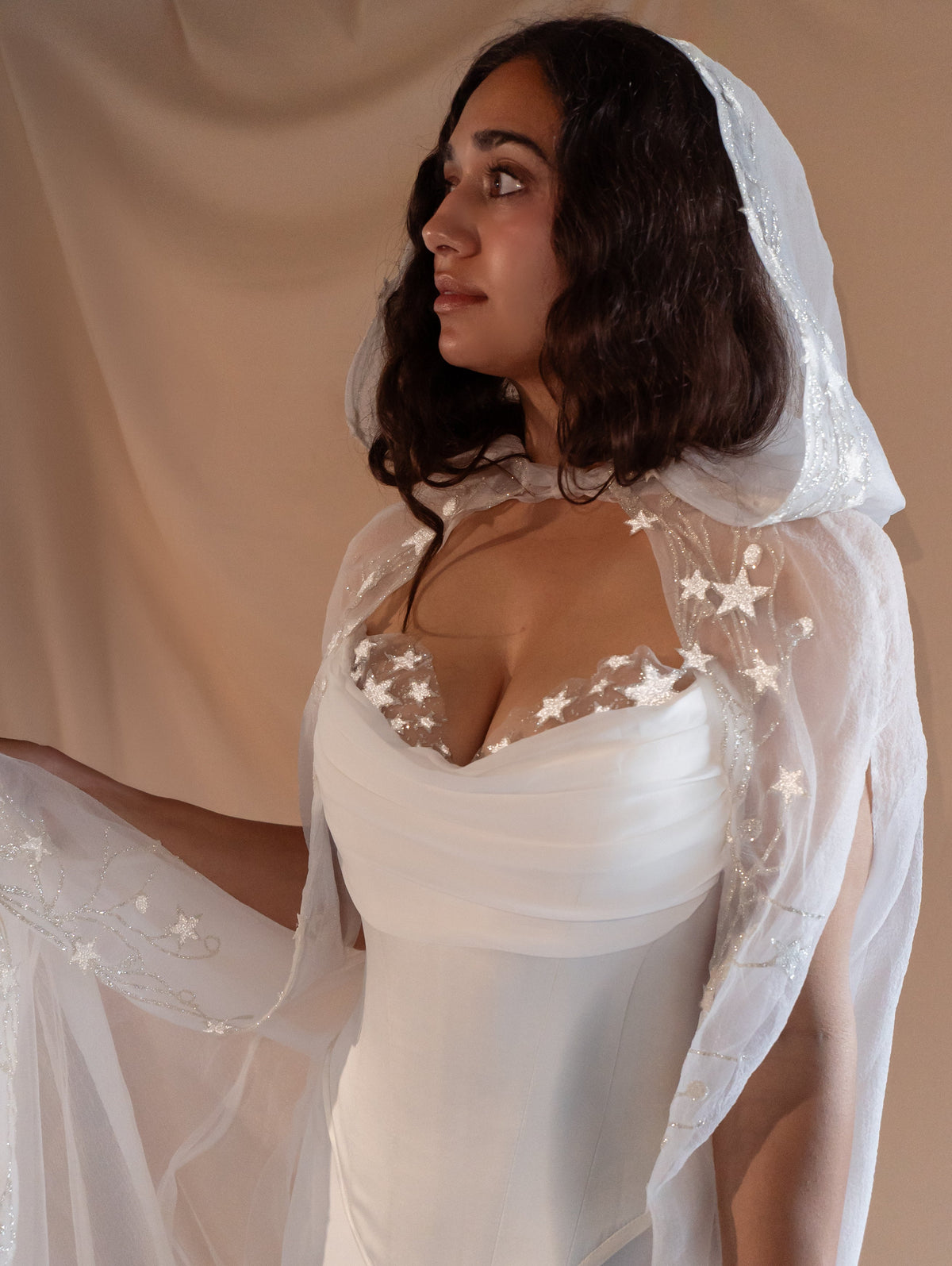 Profile of a bride in a hooded star cape over the satin corset of a bridal separates set, studio drape, Venus Ensemble