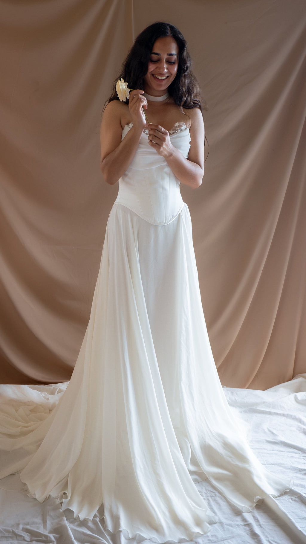 Smiling bride with a rose in a corset and skirt set, chiffon train spread across a white floor cloth, Venus Ensemble