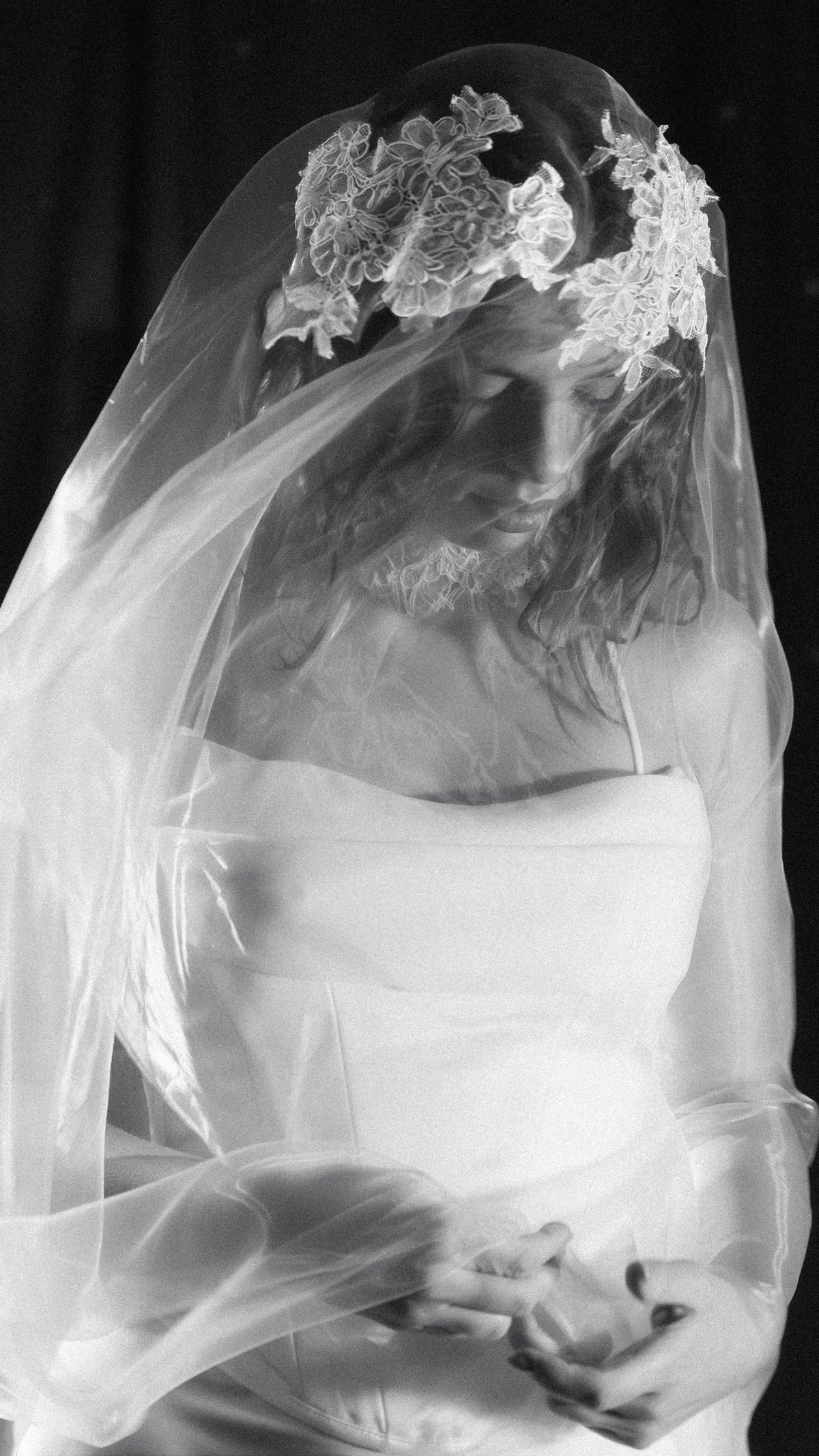 Black and white portrait of a bride looking down under a lace crown wedding veil, organza draped over her hands, mantilla veil, Vespera