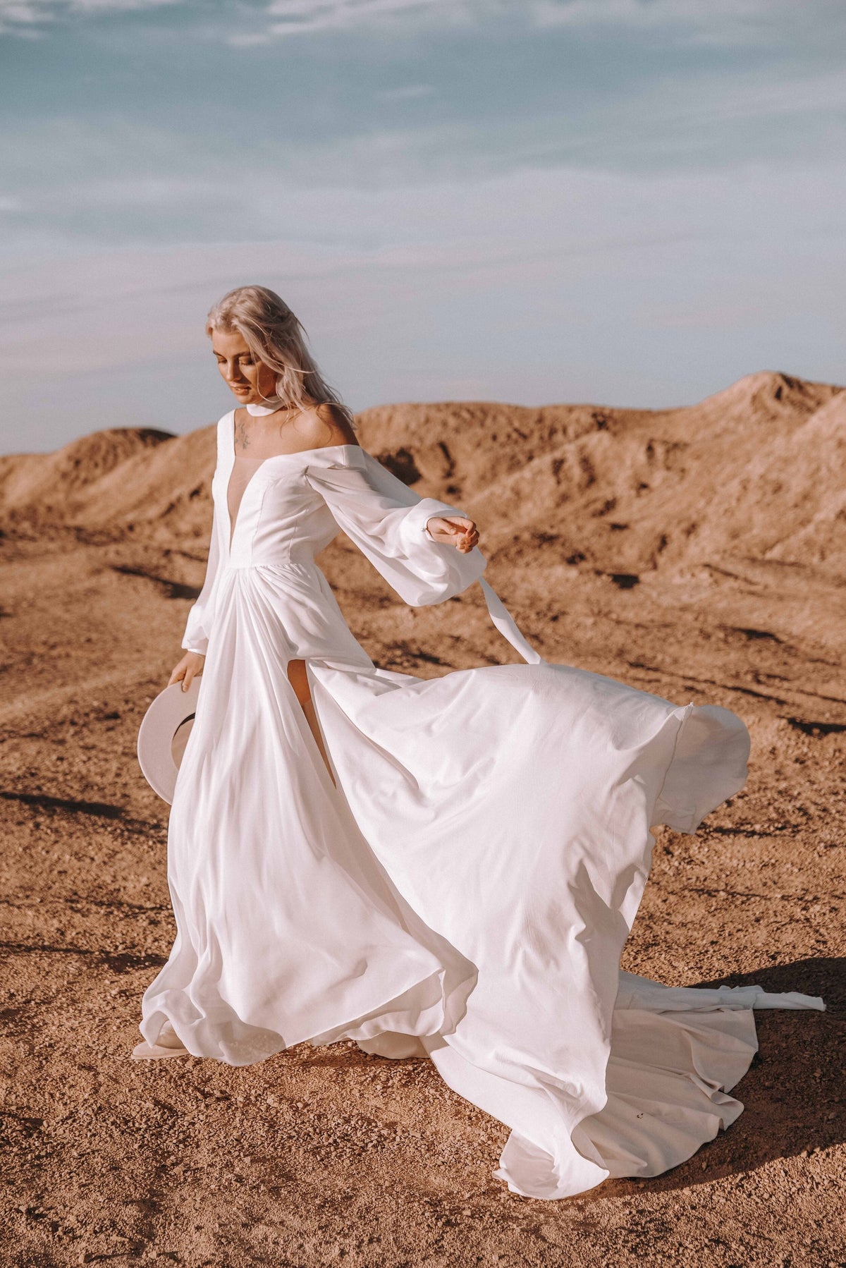 Bride twirling on red dunes in a chiffon sample wedding dress with an off-shoulder V neckline and long sleeves, hat in hand, When the Moon is Out Sample