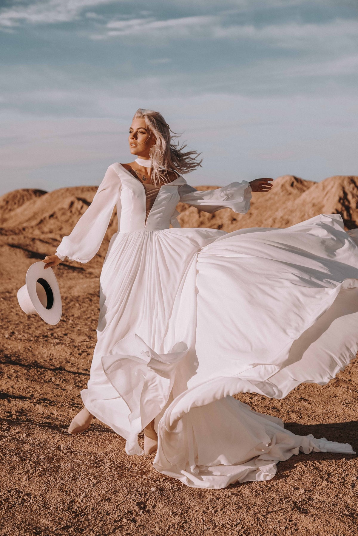 Bride flinging the chiffon skirt of a long-sleeve sample wedding dress into the wind on a desert ridge, hat in hand, When the Moon is Out Sample