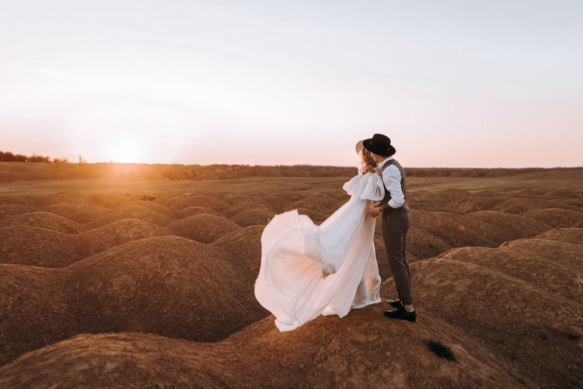 Wide view at sunset of a couple embracing on rolling hills, the bride's chiffon sample wedding dress caught by the wind, When the Moon is Out Sample