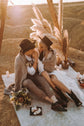 Couple sitting on a rug with pampas and candles, bride in a chiffon sample wedding dress with a knit wrap and boots, When the Moon is Out Sample
