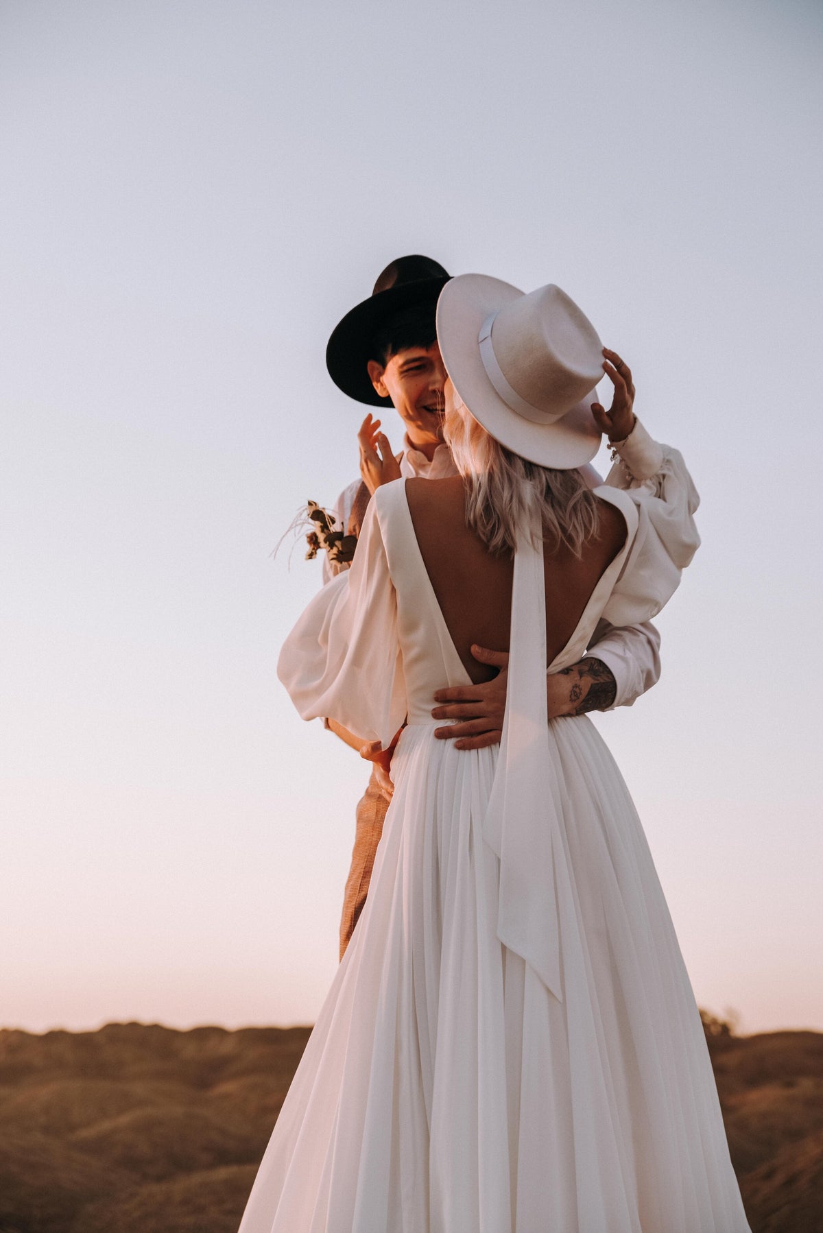 Couple embracing, bride in a hat showing the open-back chiffon sample wedding dress with the scarf trailing down, When the Moon is Out Sample