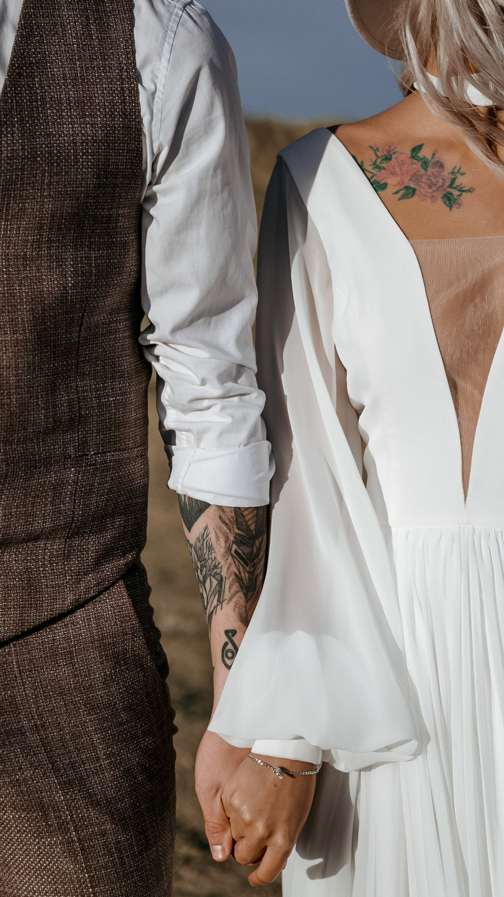 Close crop of a couple holding hands, deep V neckline and sheer sleeve of a chiffon sample wedding dress beside a tweed vest, When the Moon is Out Sample