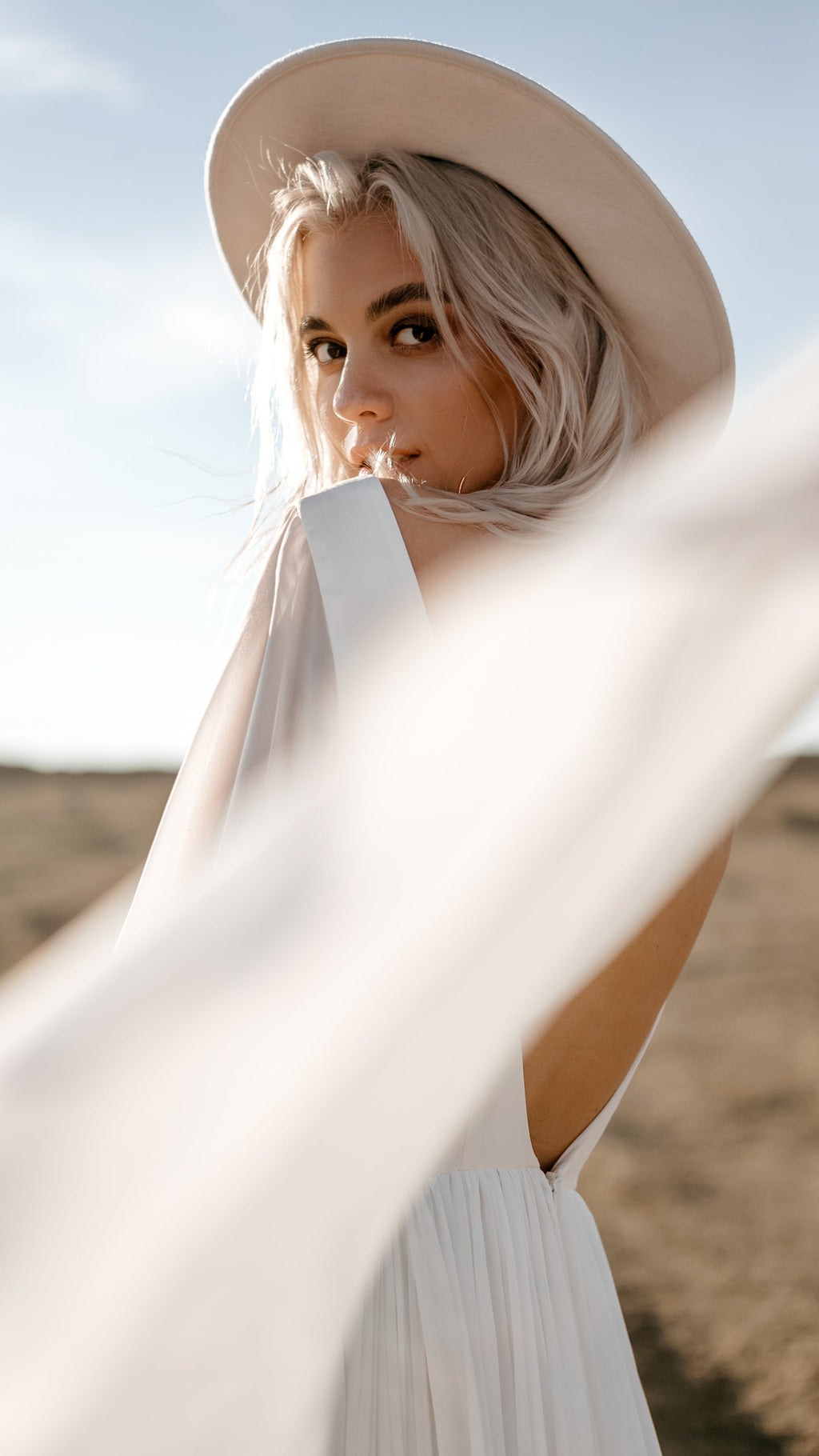 Bride in a hat looking over her shoulder, chiffon sample wedding dress sleeve streaming across the frame, open back, When the Moon is Out Sample