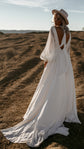 Back view of a bride in a hat on a dirt slope, keyhole back and balloon sleeves of a chiffon sample wedding dress with a train, When the Moon is Out Sample